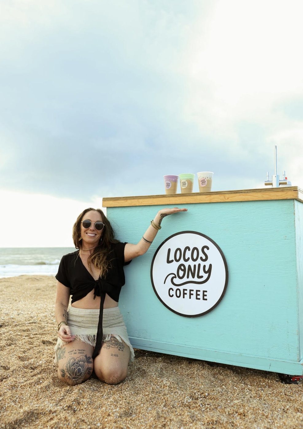 Nikki kneeling beside the turquoise Locos Only coffee cart on the beach