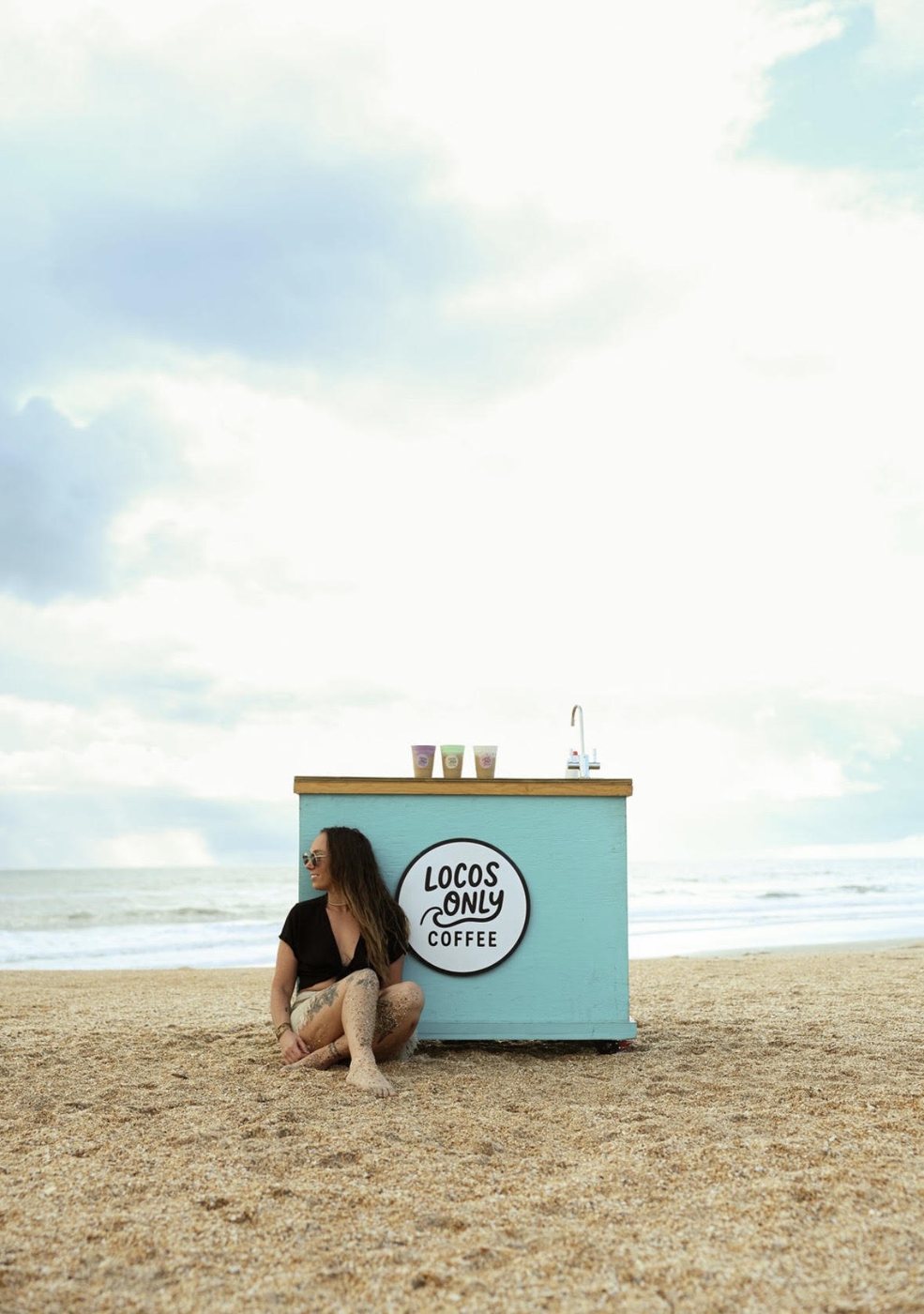 Nikki sitting in the sand against the Locos Only cart by the ocean